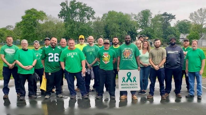 Group photo outdoors with several men in green shirts holding a banner reading “Mental Health Awareness Month.”